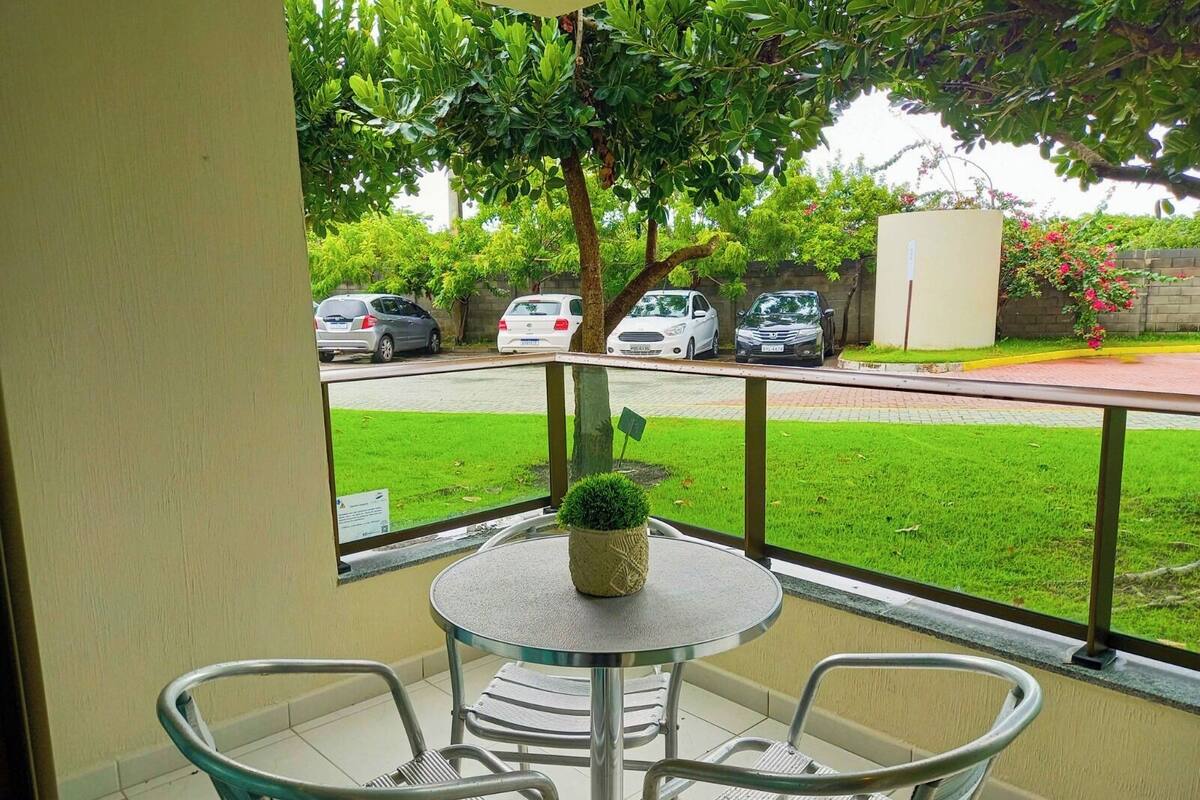 A balcony with a round table and two chairs, a potted plant, and a view of parked cars and a grassy area.