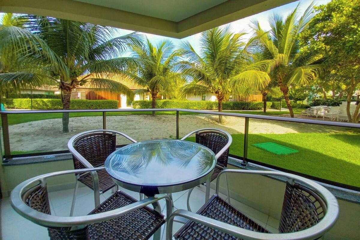A balcony with a glass table and wicker chairs, overlooking a sandy area with palm trees and a well-maintained lawn.