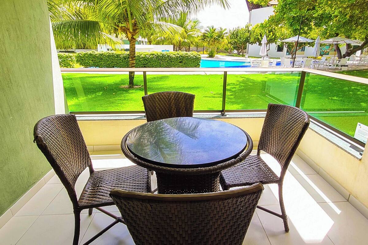 A balcony with a round table and four wicker chairs, overlooking a pool and a well-maintained lawn.