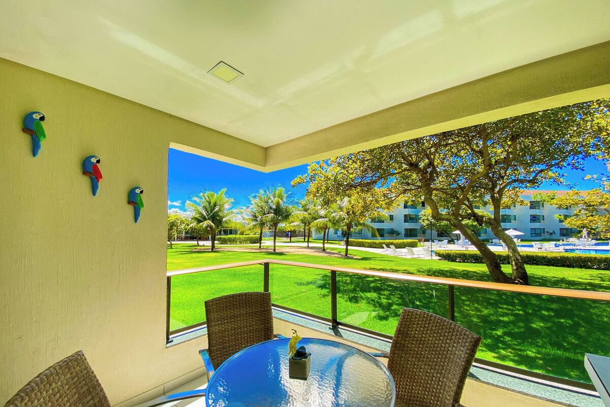 A balcony with wicker chairs, a glass table, and a view of a well-maintained lawn and buildings.