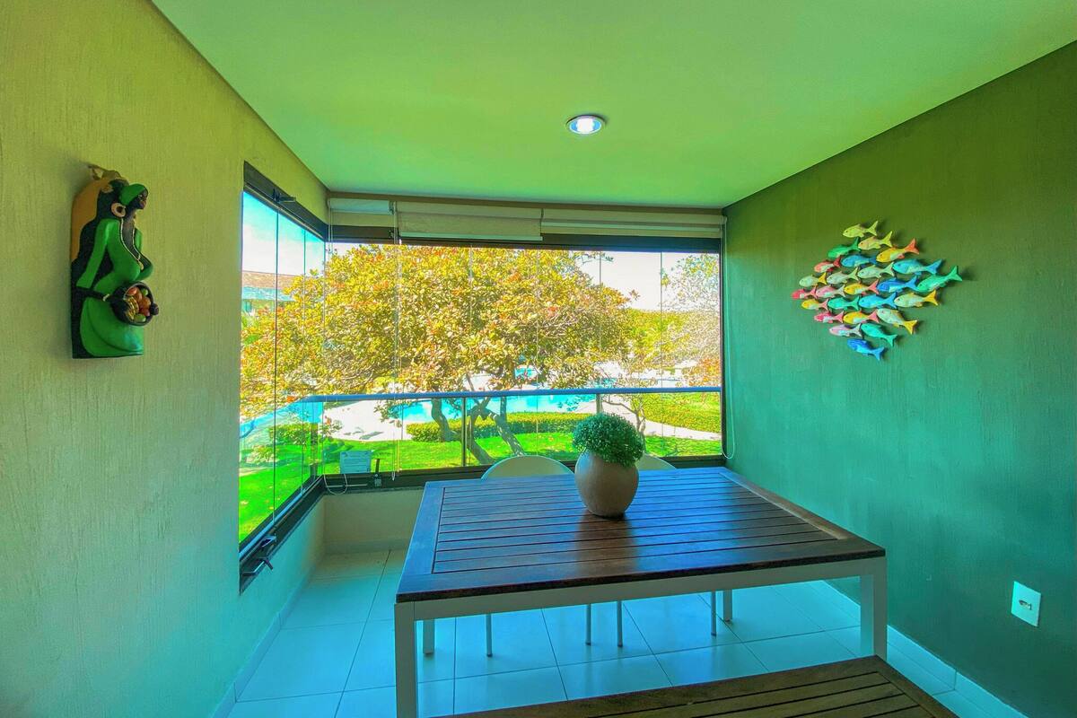 A balcony with a table and a potted plant, a green wall with a fish decoration, and a view of a tree and garden.