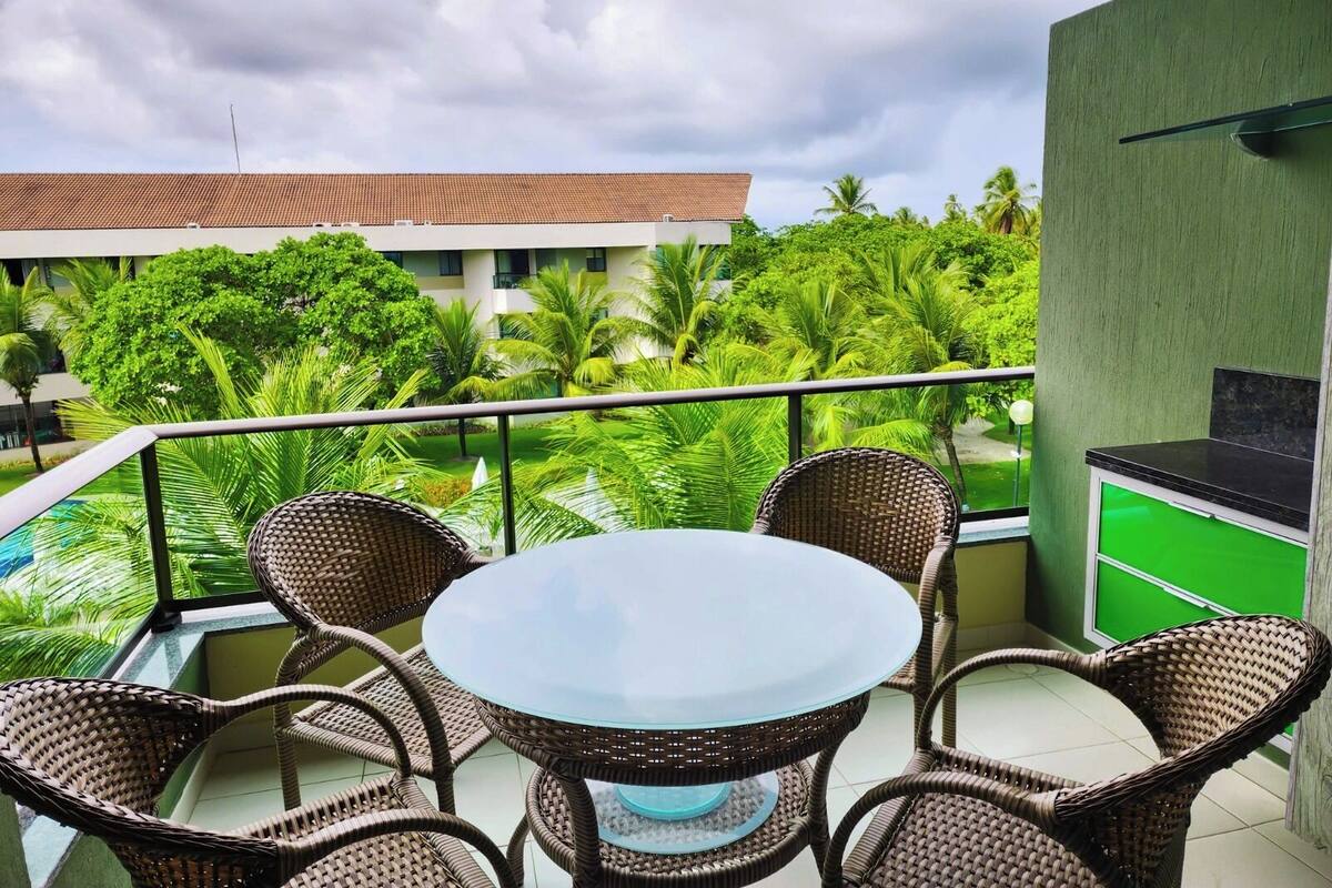 A balcony with wicker chairs and a glass table, overlooking a pool and lush greenery.