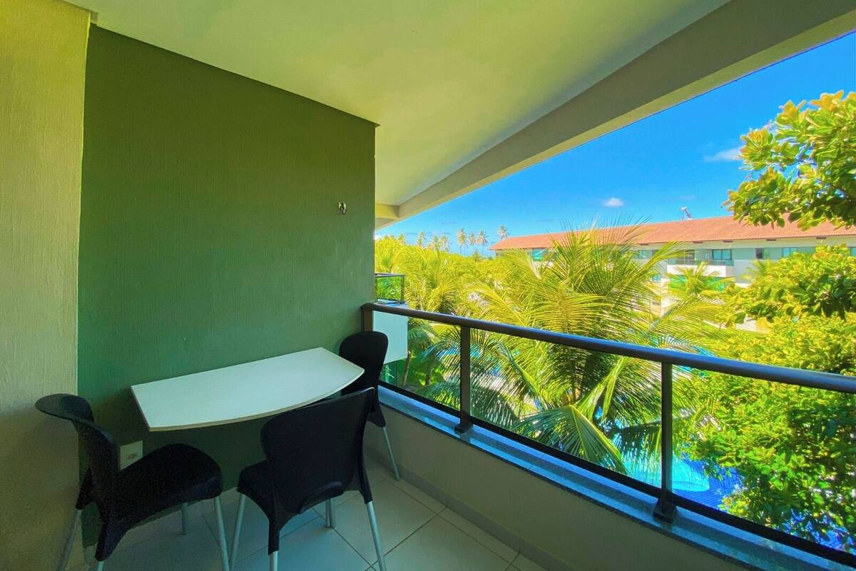 A balcony with a table and chairs, a view of a building with a red roof, and lush greenery.