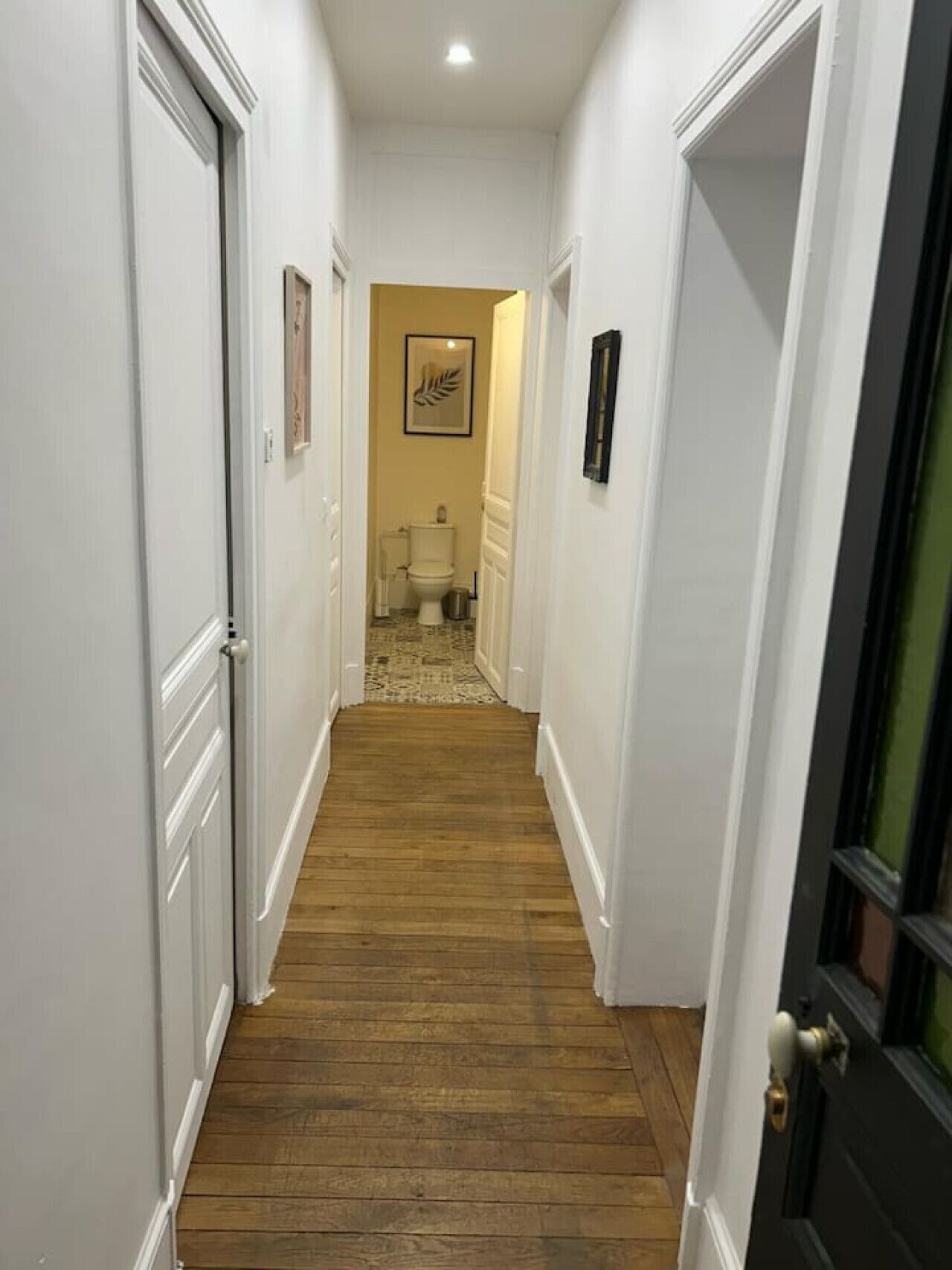 A narrow hallway with wooden flooring and white walls, leading to a bathroom with a toilet and a framed artwork on the wall.