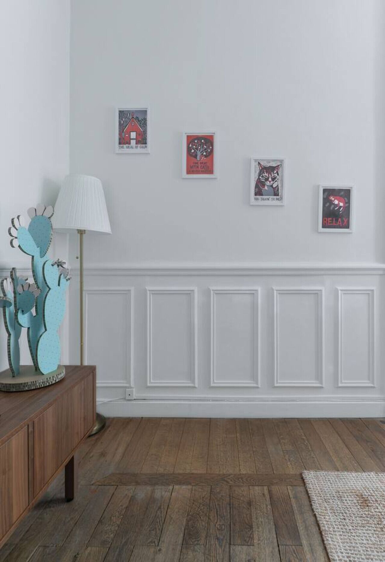 A room with a wooden floor, white wainscoting, a wooden console table with a lamp, and three framed posters on the wall.