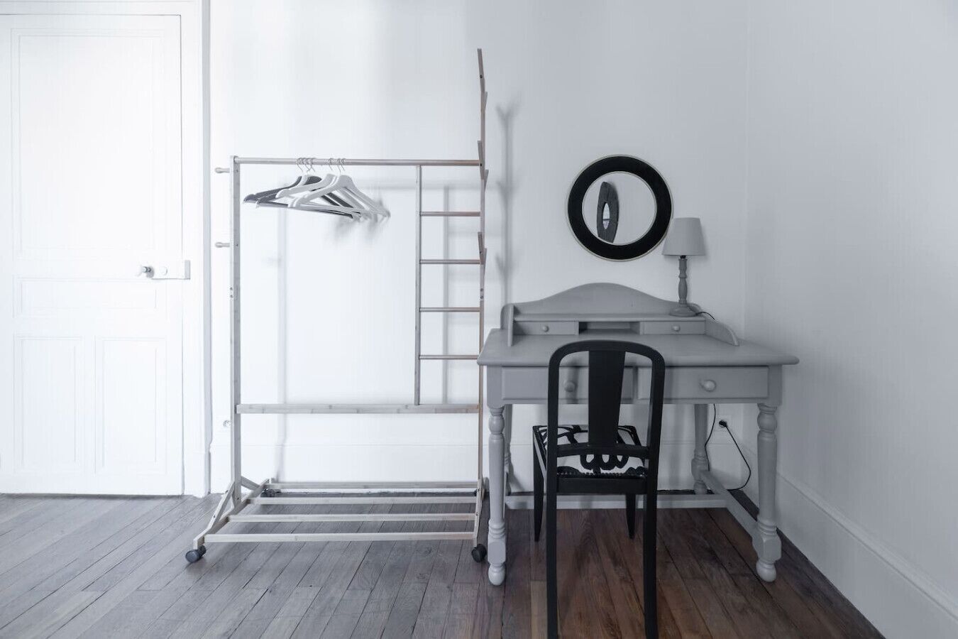 Una habitación con un escritorio blanco, una silla, un tendedero y un espejo montado en la pared.