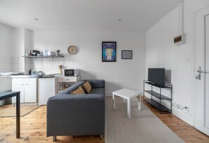 Una sala de estar moderna con un sofá gris, una mesa de centro blanca, un televisor sobre un mueble negro y una zona de cocina con armarios blancos y un microondas.