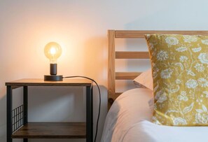 Una mesita de noche con una lámpara, una cama con ropa de cama blanca y un cojín floral, y un cabecero de madera.
