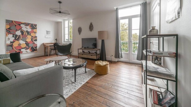 Un salon avec un canapé, une table basse et un téléviseur. Il y a une fenêtre avec des rideaux, une lampe et une bibliothèque.