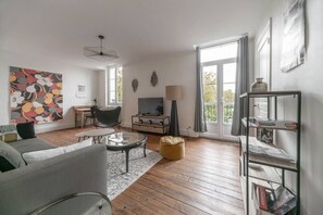 Een woonkamer met een bank, salontafel en een televisie. Er is een raam met gordijnen, een lamp en een boekenkast.