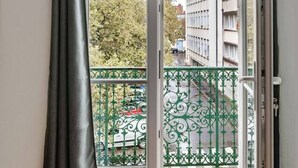 Une pièce dotée d’un plancher en bois, d’une porte-fenêtre coulissante donnant sur un balcon avec une rambarde en métal vert et offrant une vue sur un immeuble et des arbres.