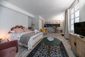 A modern bedroom with a bed, a small table, a chair, a TV, and a kitchenette in the background. - Superbe studio avec terrasse dans immeuble classé (Vichy)