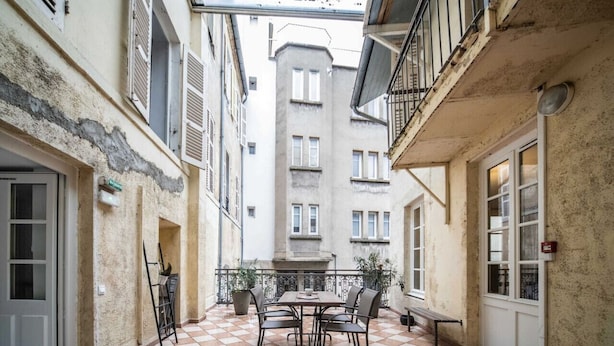 A narrow courtyard with a tiled floor, a table with chairs, and a view of a building with multiple windows. - Superbe studio avec terrasse dans immeuble classé (Vichy)
