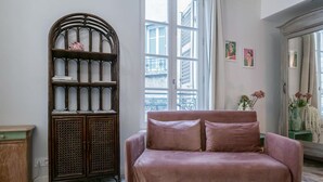 A room with a pink sofa, a wooden cabinet with glass doors, and a large window with a view of buildings. - Superbe studio avec terrasse dans immeuble classé (Vichy)