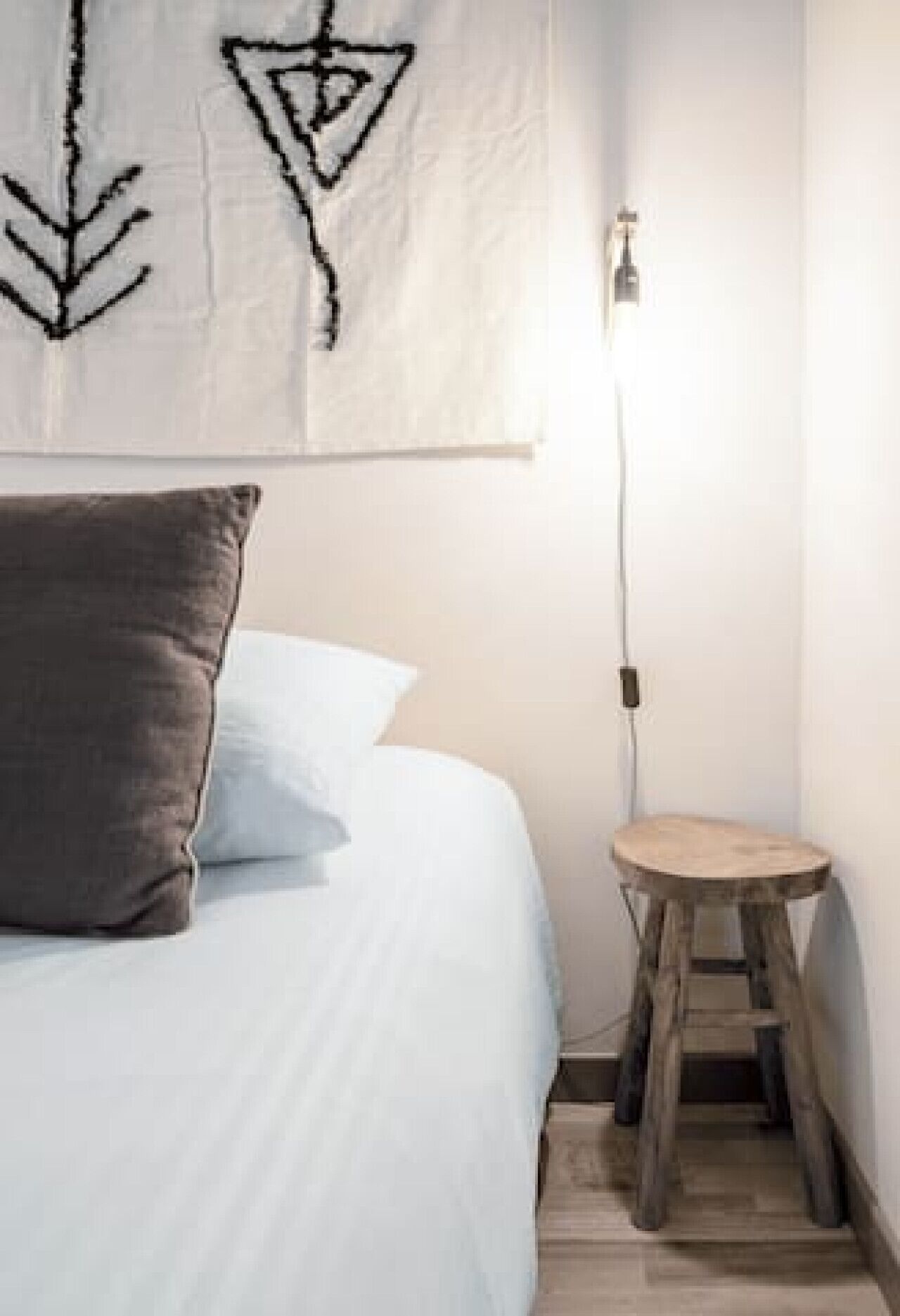 A bedroom with a bed, pillows, a small stool, and a wall-mounted light fixture.