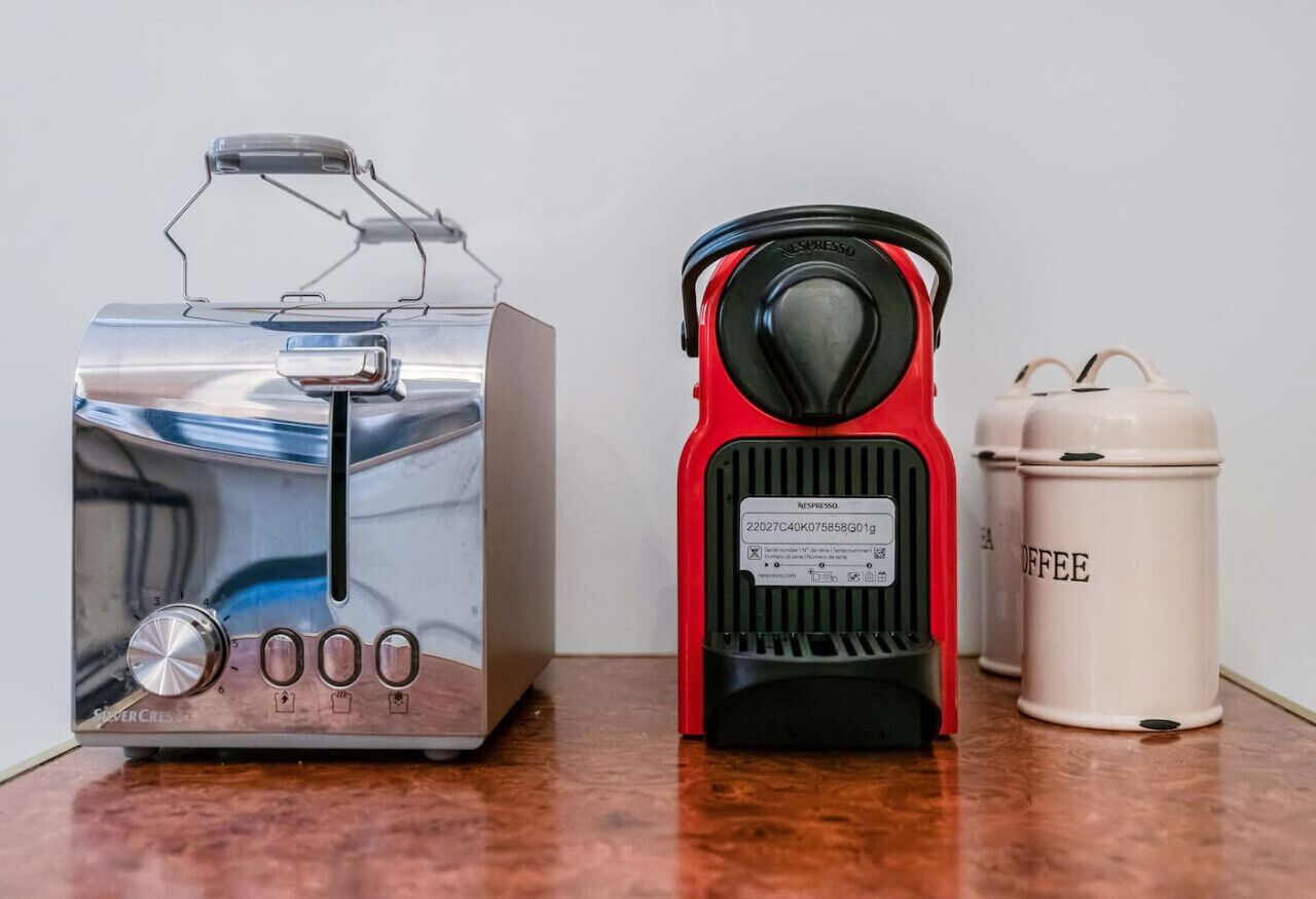 Een roestvrijstalen toaster, een rode koffiezetapparaat en een beige koffiebus op een houten oppervlak.