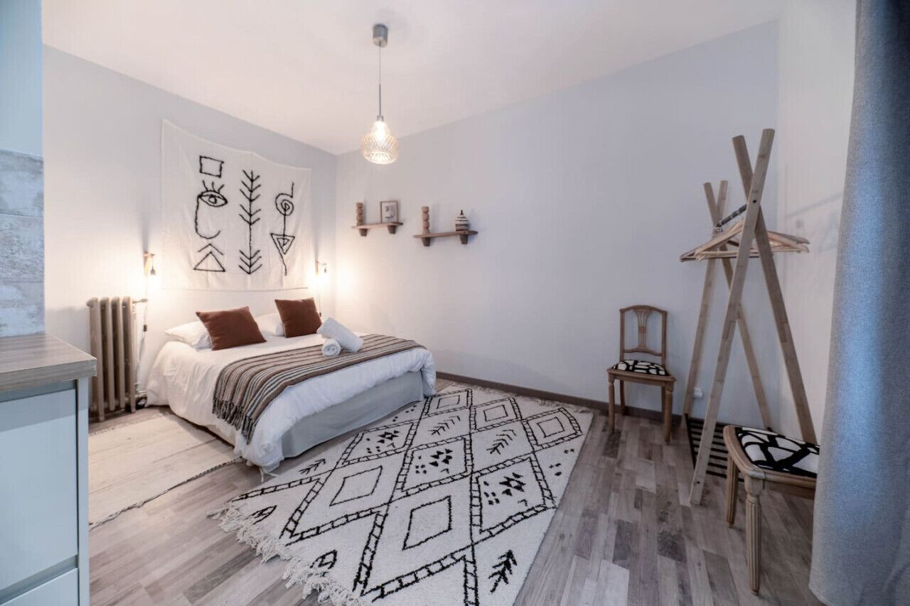 Een slaapkamer met een bed, een kamerbreed tapijt met patroon, een houten vloer, een radiator, een hanglamp, een wandtapijt en een houten schilderstatief.