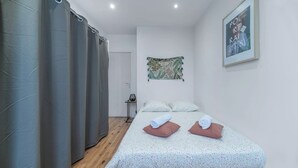 Une chambre à coucher avec un lit, deux oreillers, une petite table avec une lampe, un rideau et un tableau au mur.