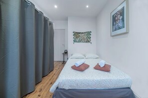 A bedroom with a bed, two pillows, a small table with a lamp, a curtain, and a painting on the wall. - Appartement Lumineux en Centre Ville Avec Parking (Vichy)