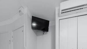 Una habitación con un armario blanco, un televisor de pantalla plana montado en la pared, una pequeña mesa redonda con una silla y suelo de madera en tonos claros.