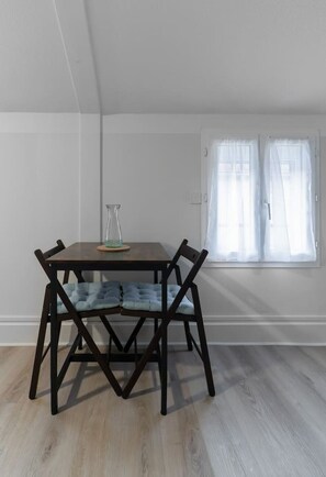 Un comedor con una mesa y sillas de madera oscura, un florero de vidrio sobre la mesa, y una ventana con cortinas translúcidas.