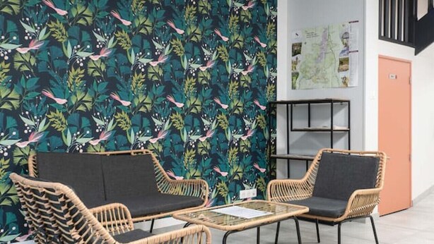 Una habitación con pared de diseño, dos sillones de mimbre con cojines negros, una mesa redonda pequeña y una alfombra tejida.