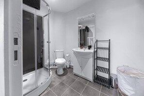 A bathroom with a glass-enclosed shower, a white toilet, a sink with a mirror, and a metal shelf unit.