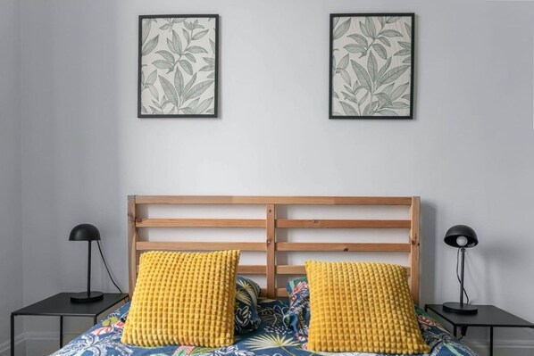 A bedroom with a wooden headboard, two bedside tables with lamps, a bed with a floral bedspread, and two framed botanical prints on the wall.