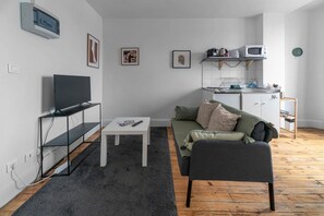 A modern living room with a flat-screen TV, a small white table, a grey sofa, and a kitchen area with a microwave.