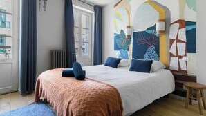 A bedroom with a bed, a blue rug, a wooden floor, a window with curtains, and a wall with abstract art. - Superbe loft climatisé dans immeuble classé (Vichy)
