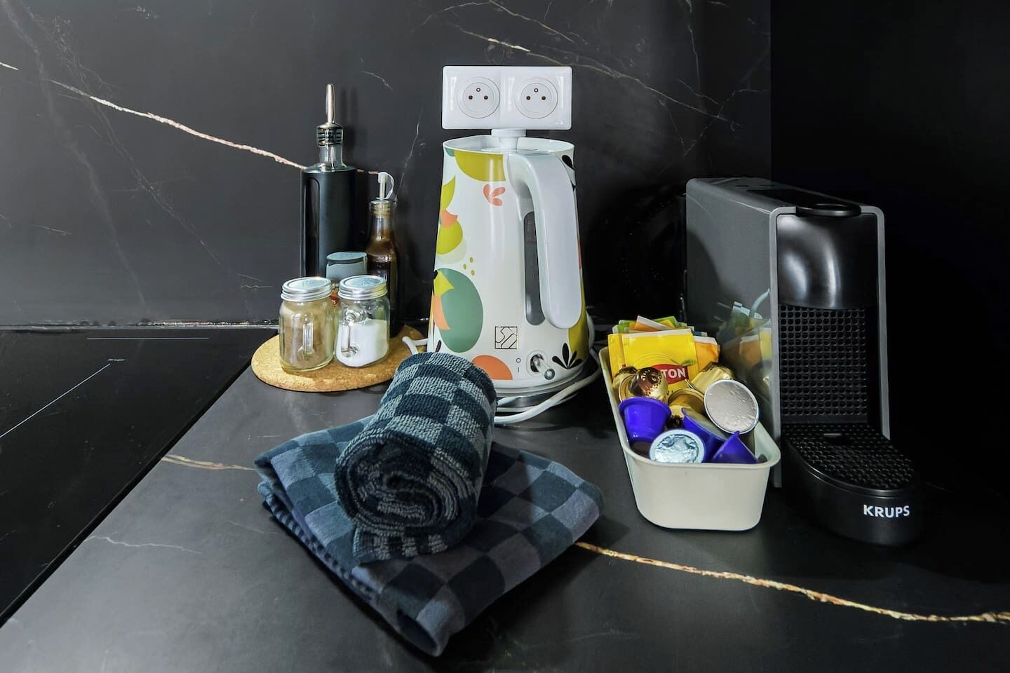 A kitchen counter with a kettle, a coffee maker, a portable heater, a container with snacks, and a rolled-up towel.