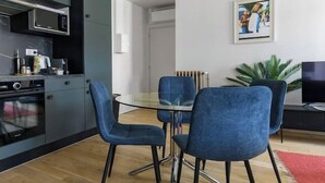 A modern kitchen with dark cabinets, a microwave, and an oven. There are two blue chairs around a glass table. A flat-screen TV is on a stand in the living area. - Superbe loft climatisé dans immeuble classé (Vichy)