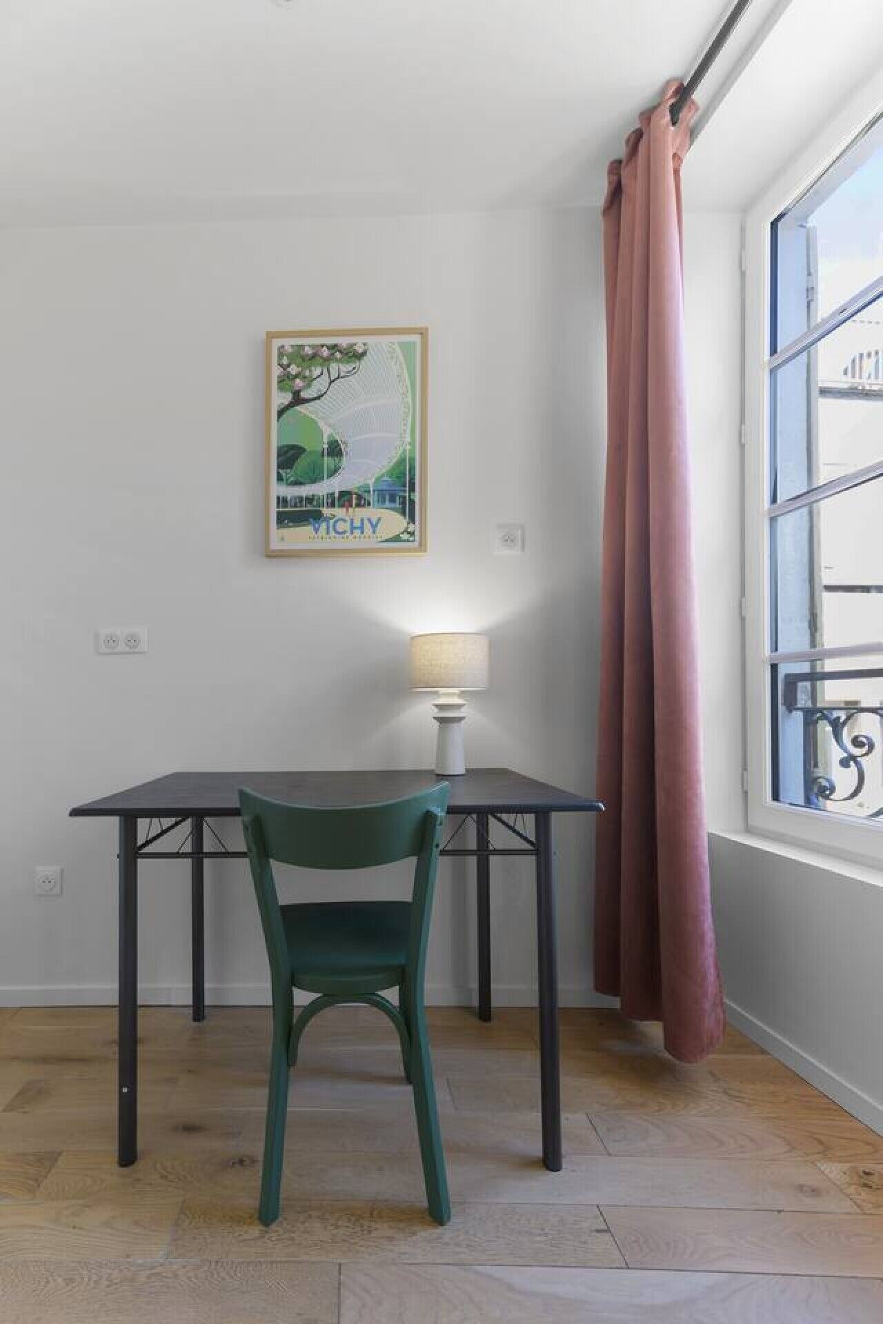 A room with a table and chair, a lamp, a framed picture on the wall, and a window with curtains.