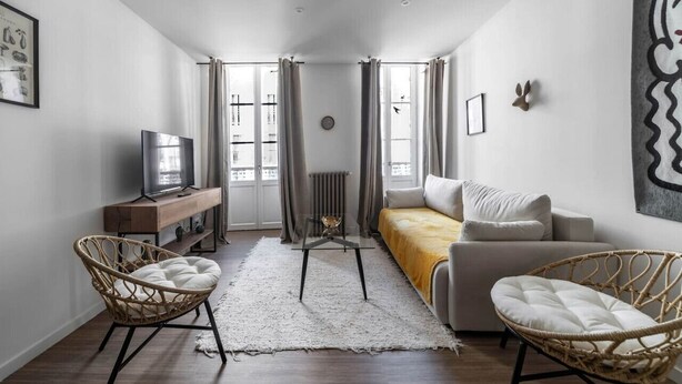 Un salon moderne comprenant un canapé, deux fauteuils en osier, une table basse en verre, un téléviseur posé sur un meuble en bois et un radiateur.