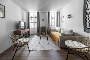 A modern living room with a sofa, two wicker chairs, a glass coffee table, a TV on a wooden stand, and a radiator. - Superbe appartement climatisé dans immeuble classé (Vichy)