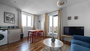 Una sala de estar moderna con un sofá azul, una mesa de comedor con sillas rojas, un televisor y una zona de cocina con lavadora.
