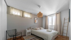 Un dormitorio con cama, una silla, una mesita de noche, una lámpara colgante, una ventana con cortinas y suelo de madera.
