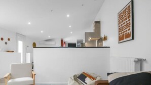 Una sala de estar moderna con un sofá, un reposapiés y un sillón, una cocina al fondo, y una obra de arte enmarcada en la pared.