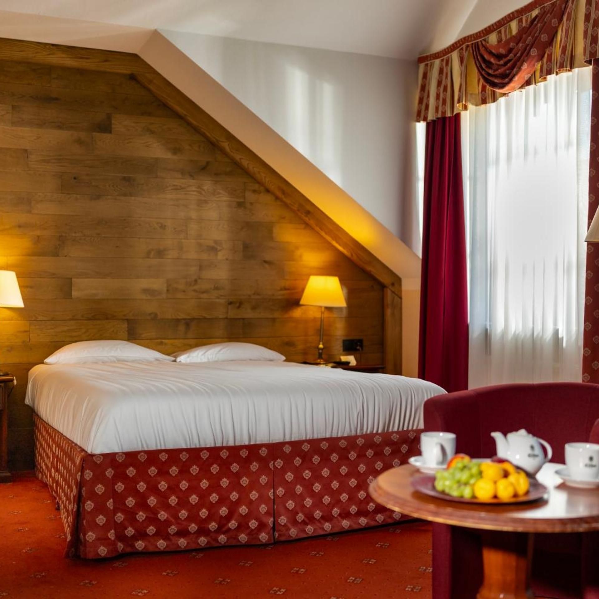 A hotel room with a wooden sloped ceiling, a bed with white linens, a red patterned skirt, a table with fruit and tea set, and a red chair.