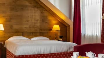 A hotel room with a wooden sloped ceiling, a bed with white linens, a red patterned skirt, a table with fruit and tea set, and a red chair.