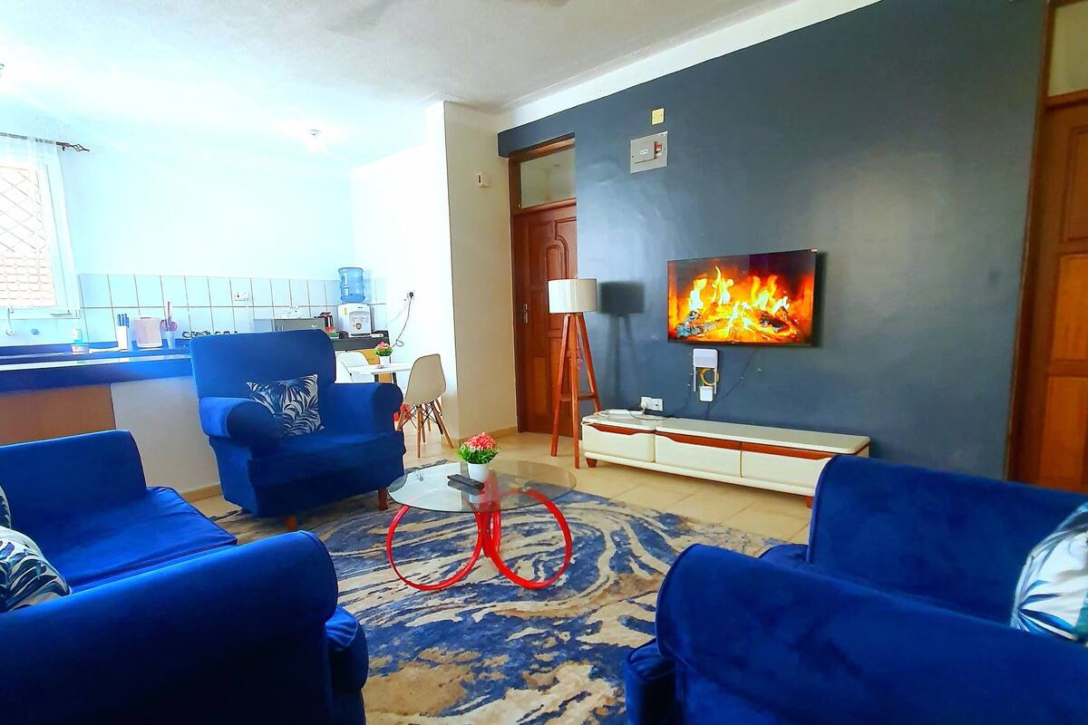 Una sala de estar con sofás azules, una mesa de centro de vidrio, una bicicleta roja, un televisor en la pared y un ventilador de techo.