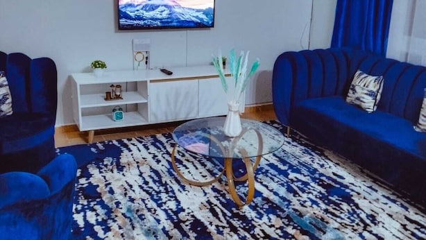 Ein Wohnzimmer mit an der Wand montiertem Fernseher, einem weißen Schrank, zwei blauen Sofas und einem gemusterten Teppich.