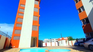 Ein Swimmingpool, umgeben von Gebäuden mit orangefarbenen und weißen Fassaden, vor einem wolkenlosen, blauen Himmel.