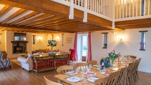 A dining area with a wooden table set for six, surrounded by wooden chairs. A fireplace is visible in the background, and the room has a high ceiling with exposed wooden beams.