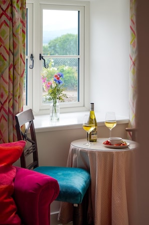 A room with a window, a chair, a table with a bottle and glass, and a vase with flowers. - Bron Haul - sleeps 2 guests in 1 bedroom (Newborough)