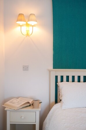 Une chambre à coucher avec un lit, une table de chevet sur laquelle se trouvent un livre et une tasse, et un luminaire fixé au mur.