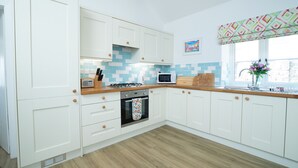 Une cuisine avec des meubles blancs, un plan de travail en bois, un four à micro-ondes, une cuisinière et une fenêtre agrémentée d’une lambrequin colorée.