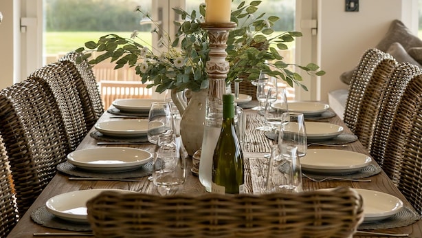 Une table à manger dressée pour six personnes, avec des chaises en osier, un vase rempli de fleurs et une bougie.