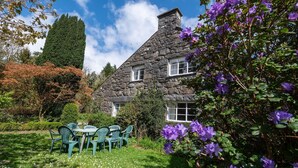 Ein Haus aus Stein mit einem geneigten Dach, umgeben von einem Garten mit violetten Blumen und grünen Sträuchern.