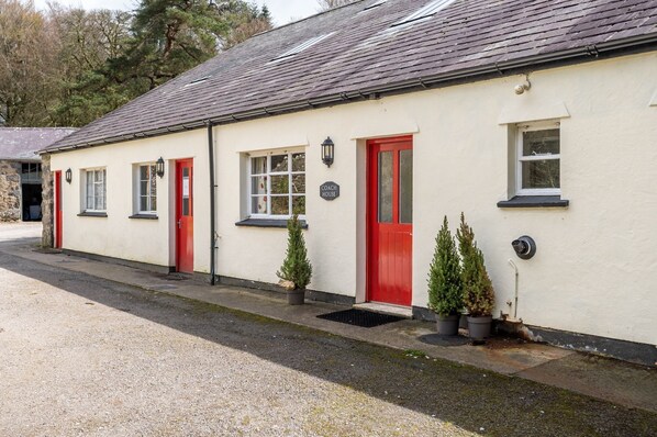 A white building with red doors and windows, labeled "COACH HOUSE," with a gravel driveway and potted plants.